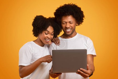 Téléchargez les photos : Joyeux milléniaux conjoints noirs couple websurfing ensemble, en utilisant un ordinateur portable sur fond de studio orange. Heureux homme et femme afro-américain regarder du contenu sur Internet - en image libre de droit