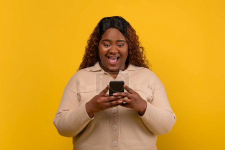 Téléchargez les photos : Heureuse dame afro-américaine millénaire dodue portant un message de lecture de chemise beige décontracté sur smartphone, ont de bonnes nouvelles, isolé sur fond de studio jaune - en image libre de droit