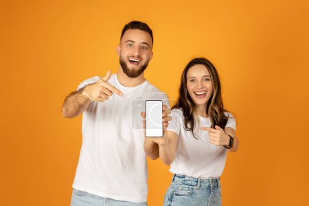 Téléchargez les photos : Jeune homme et femme caucasien excité en chemises blanches montrant un écran de smartphone vide, le pointant avec des expressions joyeuses, debout sur un fond orange vif - en image libre de droit