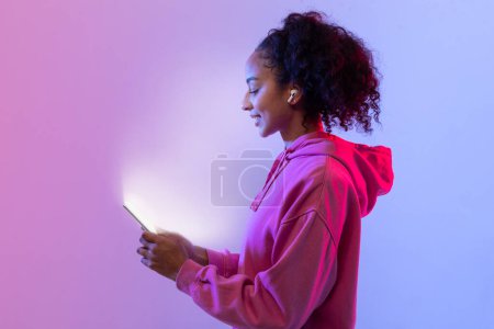Foto de Señora negra sonriendo como utiliza su teléfono inteligente con pantalla luminosa, mujer con sudadera con capucha, posando sobre fondo de neón rosa y azul, vista lateral - Imagen libre de derechos