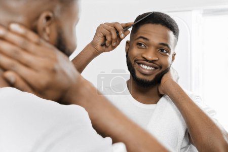 Foto de Sonriente hombre afro-americano peinando el cabello y mirando en el espejo en el baño. - Imagen libre de derechos
