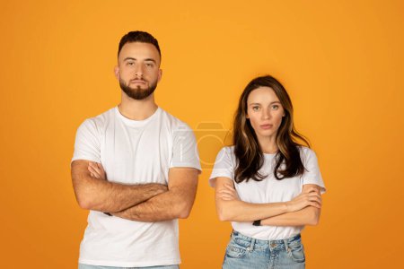 Photo for Serious and confident caucasian young man and woman in white t-shirts with arms crossed, standing with firm stances, exuding a strong and determined attitude against an orange backdrop - Royalty Free Image