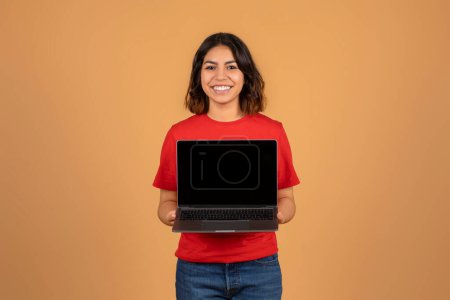 Positiv hübsche arabische Frau zeigt Laptop mit leerem Bildschirm. Lächelnde Freiberuflerin aus dem Nahen Osten, die eine Webseite empfiehlt, isoliert auf beigem Studiohintergrund. Freiberuflicher, entlegener Job
