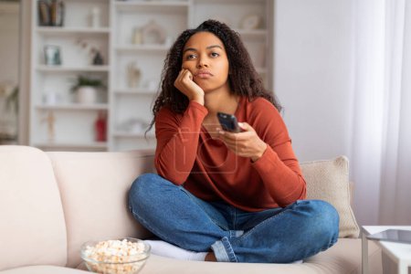 Téléchargez les photos : Jeune femme noire avec télécommande à la recherche ennuyée tout en regardant la télévision à la maison, contrarié femme afro-américaine assis sur le canapé avec bol de maïs soufflé, reflétant le mécontentement ou l'insatisfaction - en image libre de droit
