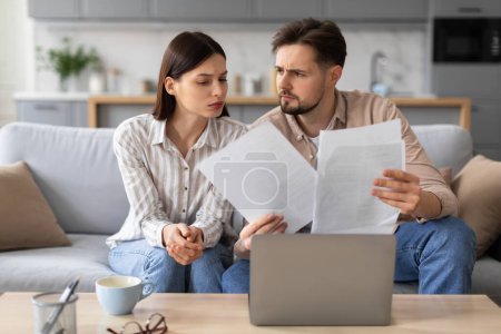Photo for Worried young couple examines paperwork together, sitting on couch in front of open laptop and with cup of coffee on table, in living room home setting - Royalty Free Image