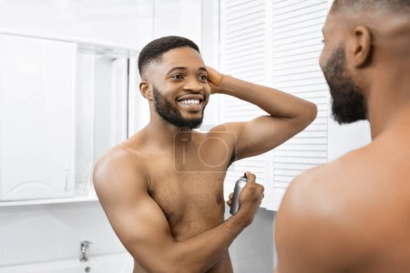Téléchargez les photos : Homme afro souriant avec torse musculaire nu à l'aide de spray corporel, regardant dans le miroir dans la salle de bain. Mans concept de soins quotidiens - en image libre de droit