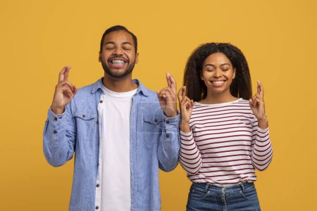 Photo for Cheerful Young Black Couple Crossing Fingers, Making Wish With Closed Eyes, Positive African American Man And Woman Pleading For Luck And Fortune, Standing Together Against Yellow Studio Background - Royalty Free Image
