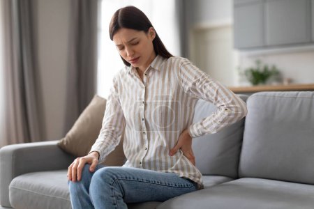 Concerned young woman sitting on couch, clutching her lower back with pained expression, possibly experiencing discomfort or back spasm