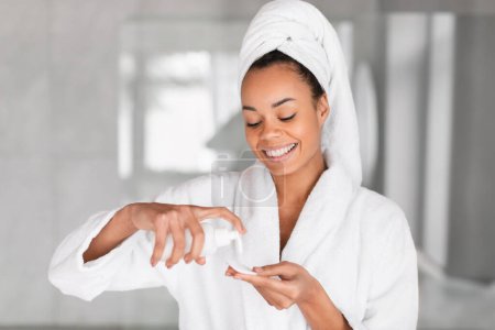 Photo for Happy African American woman with wrapped towel on head applying tonic on cotton pad, caring for facial skin with radiant smile while standing in home bathroom indoors. Skincare products - Royalty Free Image
