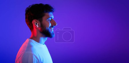 Photo for Profile portrait of smiling young man wearing wireless earphones enjoying digital music on his player, posing with eyes closed in purple blue neon light on studio background. Panorama, free space - Royalty Free Image