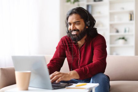 Photo for Cheerful millennial indian man working from home, sitting on couch in living room, using headset and laptop computer, have online meeting with colleagues, copy space - Royalty Free Image