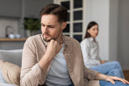 Focused man pondering deeply on foreground with contemplative expression, as blurred woman sits on background, suggesting tension, having relationship problems
