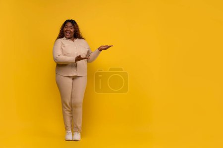Téléchargez les photos : Femme afro-américaine millénaire potelée positive portant un costume beige montrant l'espace de copie vierge pour la publicité, isolée sur fond de studio jaune, pleine longueur - en image libre de droit