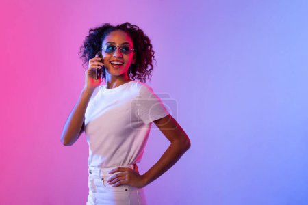 Photo for Fashionable black woman with curly hair chatting on cellphone, confidently posing in white attire with reflective sunglasses against neon-lit background, copy space - Royalty Free Image