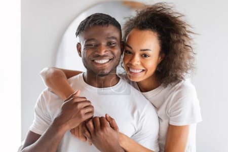 Téléchargez les photos : Portrait de heureux jeune couple noir vêtu de t-shirts blancs, étreint dans le cadre de la salle de bain, souriant à la caméra, symbolisant le lien d'amour et des moments partagés d'autosoin. choyer la famille - en image libre de droit