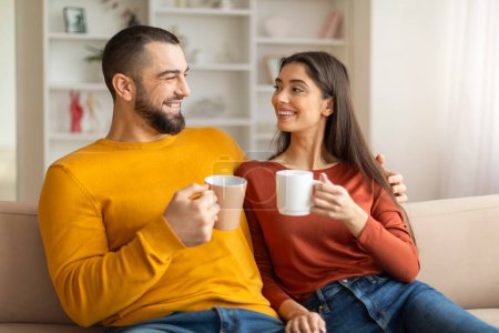 Photo for Young couple in love drinking coffee together, sitting on couch in living room, happy caucasian man and woman in casual clothes enjoying hot drink at home, chatting and smiling, free space - Royalty Free Image