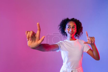 Photo for Vibrant young black woman with curly hair excitedly interacts with virtual touch screen, fingers extended, against dynamic pink and blue neon backdrop - Royalty Free Image