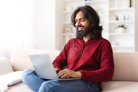 Photo for Positive millennial bearded eastern man sitting on couch, typing on computer laptop keyboard, living room interior, distant employee working from home, copy space - Royalty Free Image
