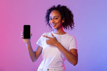 Téléchargez les photos : Femme noire joyeuse avec des cheveux bouclés pointant vers l'écran vierge du smartphone, présentant une nouvelle application sur fond lumineux néon rose et bleu vibrant - en image libre de droit