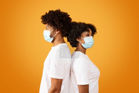 Photo for African american man and woman wearing protective medical face masks standing back to back, isolated on orange studio background. Healthcare, season cold, flu, influenza - Royalty Free Image