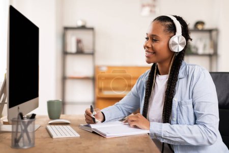 Photo for Happy black female student wearing headphones engages in e-learning, smiling as taking detailed notes from informative online lecture at her desk at home - Royalty Free Image