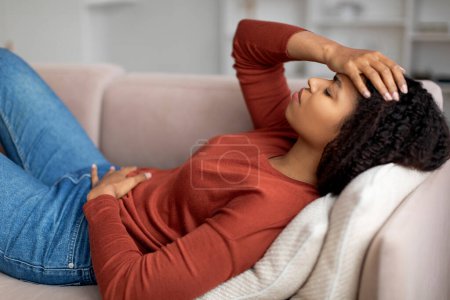Téléchargez les photos : Malade femme noire couchée sur le canapé, mal à l'aise à la maison, dame afro-américaine malade souffrant de douleurs menstruelles, contrariée femme ayant des douleurs abdominales et des maux de tête aigus, vue de côté - en image libre de droit