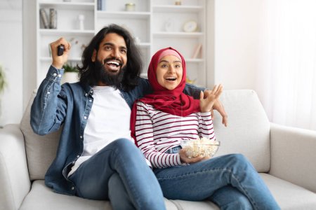 Téléchargez les photos : Joyeux jeune couple musulman relaxant sur le canapé avec du pop-corn, ayant soirée cinéma à la maison, heureux excité arabe homme et femme dans le hijab émotionnellement réagir au divertissement sur leur écran de télévision - en image libre de droit