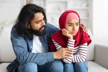 Téléchargez les photos : Homme arabe bienveillant réconfortant sa femme musulmane bouleversée à la maison, s'excusant après la querelle, offensé dame islamique dans le hijab assis sur le canapé dans le salon, jeune couple souffrant crise relationnelle - en image libre de droit