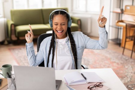 Photo for Energetic black teen female student enjoying break, dancing with headphones on while working at her desk filled with notes and laptop in bright room - Royalty Free Image