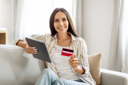 Photo for Beautiful Young Woman With Digital Tablet And Credit Card In Hands Sitting On Couch At Home, Happy Smiling Millennial Female Enjoying Online Shopping And Easy Payments, Looking At Camera, Copy Space - Royalty Free Image