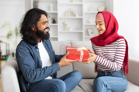 Foto de Hombre árabe dando regalo a su esposa musulmana emocionada en casa, sorprendente cónyuge feliz con caja de regalo mientras se sientan en el sofá juntos, saludo con San Valentín o Día Internacional de la Mujer, primer plano - Imagen libre de derechos