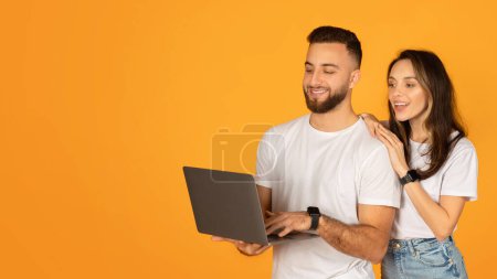 Photo for Smiling young man holding a laptop with attentive woman by his side, both wearing white t-shirts, seemingly engaged in an interesting online discovery, against a vivid orange background - Royalty Free Image