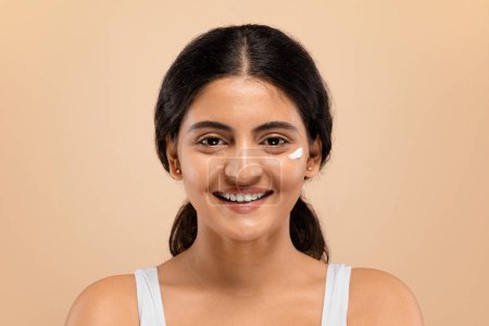 Foto de Cuidado de la piel. Mujer india joven con una pizca de crema facial en su mejilla sonriendo a la cámara, hermosa hembra oriental de pie sobre un fondo beige, destacando la rutina diaria de cuidado de la piel, espacio libre - Imagen libre de derechos