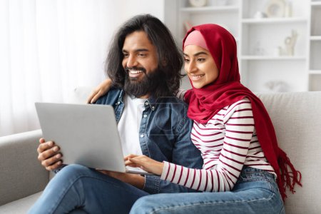 Foto de Retrato de cónyuges musulmanes felices relajándose con el ordenador portátil en casa, sonriendo joven pareja árabe abrazando y utilizando la computadora juntos, Descansando en el sofá en la sala de estar, navegar por Internet o ir de compras en línea - Imagen libre de derechos