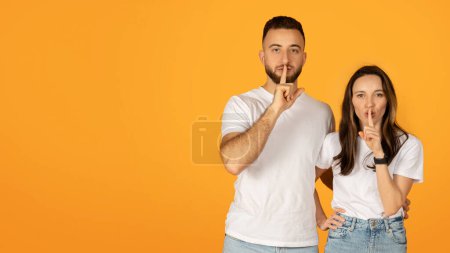 Quietly confident man and woman in white shirts placing fingers on lips in a shush gesture, suggesting secrecy or silence, standing side by side with a bright orange background