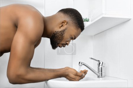 Photo for Young afro man with bare torso washing face in bathroom. Morning hygiene concept, copy space - Royalty Free Image