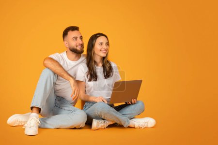 Téléchargez les photos : Couple caucasien détendu et heureux en hauts blancs et jeans bleus assis jambes croisées sur le sol tout en partageant un ordinateur portable, regardant vers le côté avec intérêt sur un fond orange - en image libre de droit