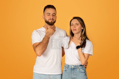Jeune homme et jeune femme caucasiens réfléchis touchant leur gorge, les yeux fermés, indiquant des sensations de douleur ou d'inconfort, portant des t-shirts blancs unis sur un fond orange