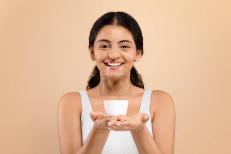 Téléchargez les photos : Meilleurs cosmétiques. Belle jeune femme indienne tenant pot avec crème hydratante et souriant à la caméra, attrayant Millennial femelle en blanc Top montrant un produit de beauté pour la peau, Copier l'espace - en image libre de droit