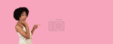 Foto de Sorprendida mujer afroamericana milenaria con la mano en la mejilla apuntando hacia los lados, expresando sorpresa e incredulidad, en un vestido floral sobre un fondo rosa, estudio, panorama - Imagen libre de derechos