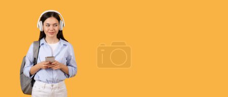 Foto de Estudiante joven contemplativa con auriculares sostiene teléfono inteligente, mirando a un lado en el espacio libre con expresión reflexiva, telón de fondo amarillo, pancarta - Imagen libre de derechos