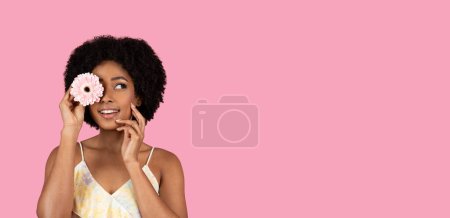Foto de Juguetona mujer afroamericana milenaria con un peinado natural sosteniendo una gerbera rosa sobre un ojo y sonriendo, creando un momento caprichoso sobre un telón de fondo rosa, estudio, panorama - Imagen libre de derechos