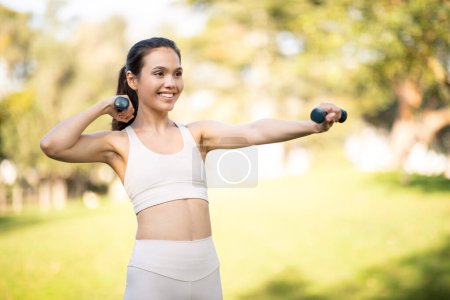 Téléchargez les photos : Une jeune femme caucasienne concentrée en tenue de fitness étend son bras avec un haltère, incarnant la force et la détermination dans un parc luxuriant. Sport, fitness, perte de poids et puissance - en image libre de droit