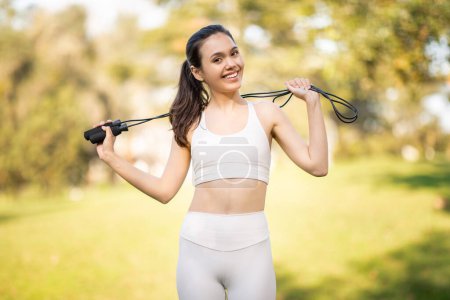 Téléchargez les photos : Une jeune athlète caucasienne joyeuse dans un soutien-gorge de sport blanc et leggings sourit vivement tout en tenant une corde à sauter sur ses épaules dans un cadre de parc luxuriant, profiter de l'entraînement cardio, le sport - en image libre de droit