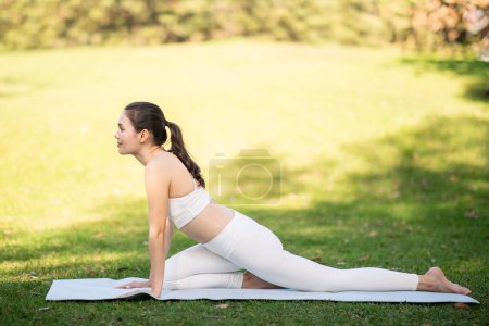 A relaxed smiling caucasian young woman athlete in all-white activewear gently stretches in a pigeon yoga pose on a mat amidst the greenery of a sunlit park, outside, full length