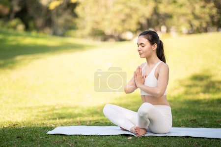 Foto de Una joven atleta caucásica serena en traje de yoga blanco medita con los ojos cerrados, las manos en oración, en una esterilla en un parque iluminado por el sol, afuera, de cuerpo entero. Zen, en forma, atención médica - Imagen libre de derechos