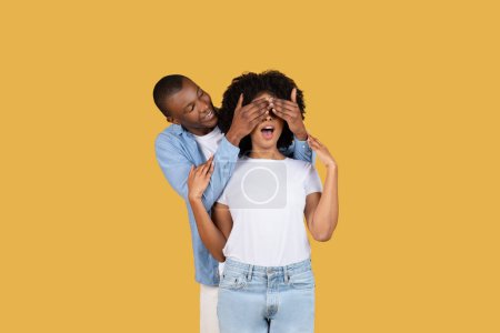 Téléchargez les photos : Un homme afro-américain millénaire ludique surprend une femme en se couvrant les yeux par derrière, souriant et debout sur un fond jaune solide, studio. Romantique date émotions - en image libre de droit