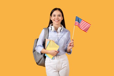 Téléchargez les photos : Étudiante rayonnante portant des cahiers et des écouteurs sur son cou, tient le drapeau américain, incarnant l'optimisme et la poursuite des connaissances sur fond jaune vif - en image libre de droit