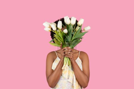 Photo for Unseen millennial African American woman standing behind a large bouquet of fresh white tulips, with just her eyes visible, against a plain pink background, studio. Holiday celebration - Royalty Free Image