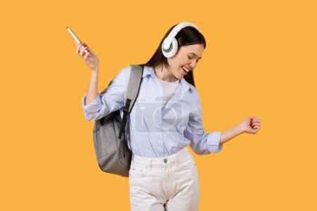 Photo for Cheerful woman student with headphones is moment of joy, dancing while holding smartphone against vibrant yellow background, embodying freedom and happiness - Royalty Free Image
